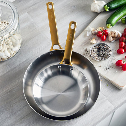 Stainless steel frying pans with gold handles on a kitchen counter with vegetables and spices.