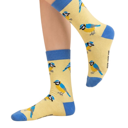 A pair of crew socks worn on a person's feet, featuring a repeating pattern of blue tits on a pale yellow background with blue ribbed cuffs and blue toes and heels.