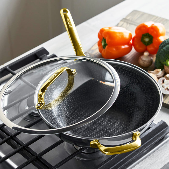 Gold ceramic wok with lid on a stove, next to fresh peppers, mushrooms, and broccoli.