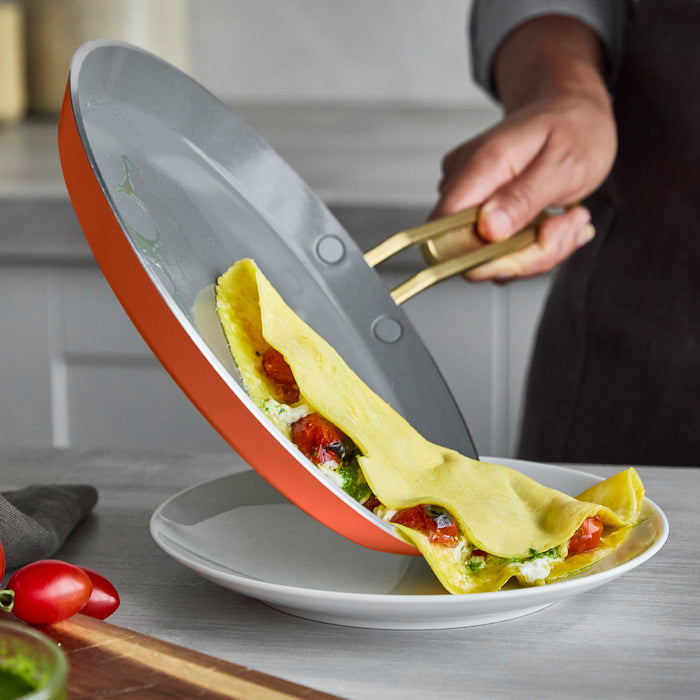 A folded omelet with tomatoes and greens being slid from a Stanley Tucci 10 nonstick pan onto a plate.