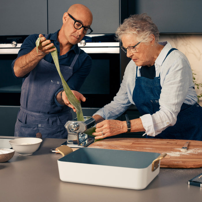 Two people roll green pasta dough beside a Stanley Tucci lasagna pan on a kitchen counter.