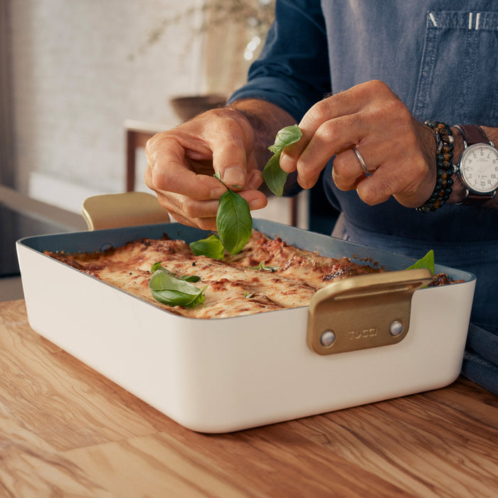 Baked pasta being garnished in a white ceramic lasagna pan on a table.