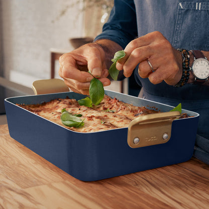 Baked dish being garnished in a blue Stanley Tucci ceramic lasagna pan on a wooden table.