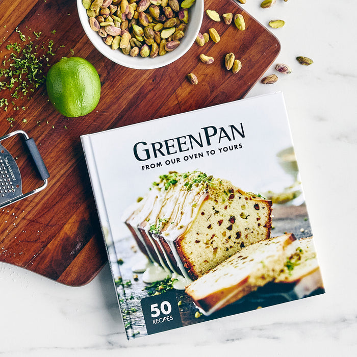 Baking cookbook on a wooden cutting board with pistachios, a lime, grater, and recipe pages nearby.