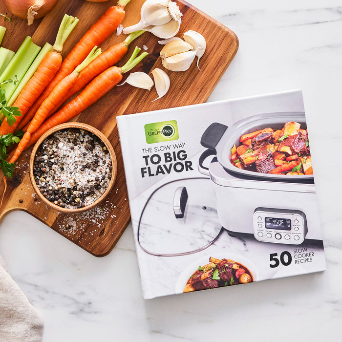 A cookbook titled The Slow Way to Big Flavor sits beside a wooden board with veggies and seasoning, its cover showcasing a stew made in the Elite 14-in-1 6-Quart Slow Cooker | Sage—ideal for fans of easy presets and advanced cooking.