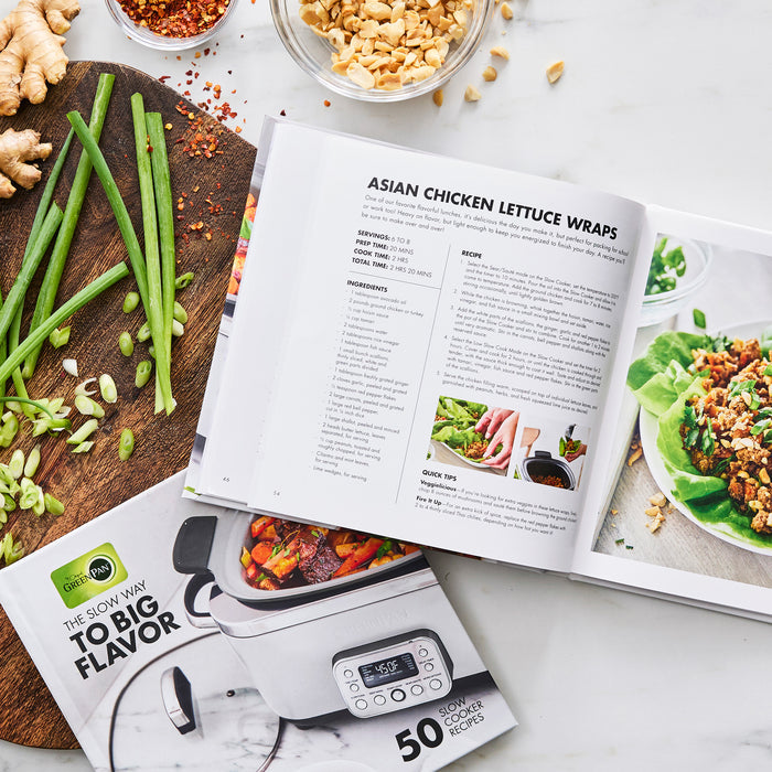An open cookbook displays a recipe for Asian chicken lettuce wraps, surrounded by green onions, ginger, crushed peanuts, and The Slow Way to Big Flavor Slow Cooker Cookbook, Vol. 2 on a white surface.