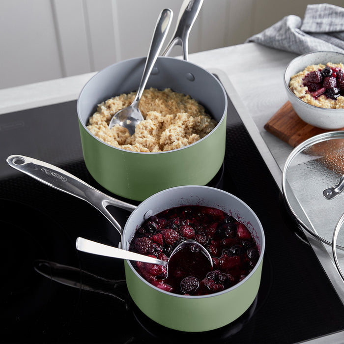 Two sage green ceramic nonstick saucepans with spoons, one filled with couscous, one with berry compote, on a stove.