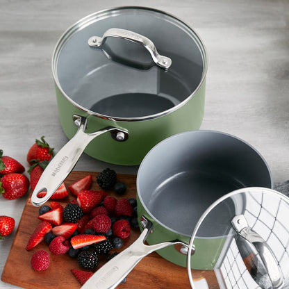 Set of two sage green ceramic saucepans with lids next to fresh berries on a board.