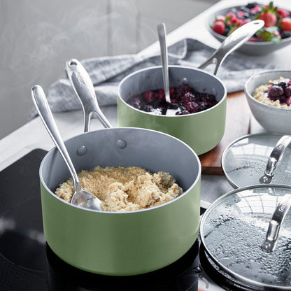 Two sage green ceramic saucepans with lids on a stove, one filled with oatmeal topped with berries. Set of two sizes.