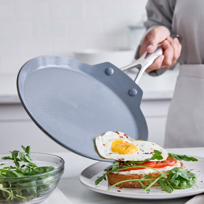 Fried egg being slid from a ceramic nonstick crepe pan onto toast with tomato and greens.
