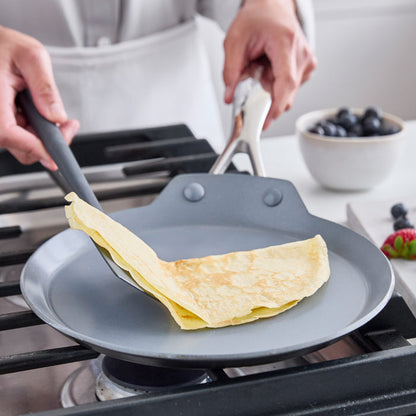 Crepe being flipped in a 9.5-inch ceramic nonstick pan; bowl of blueberries visible in the background.