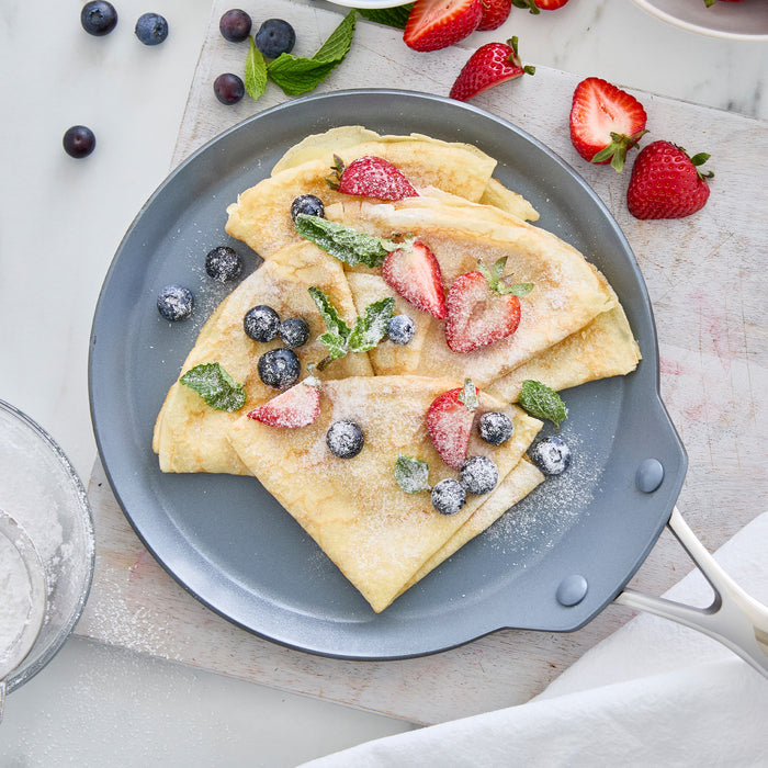 Crepes with powdered sugar, fresh berries, and mint served in a black ceramic nonstick crepe pan.