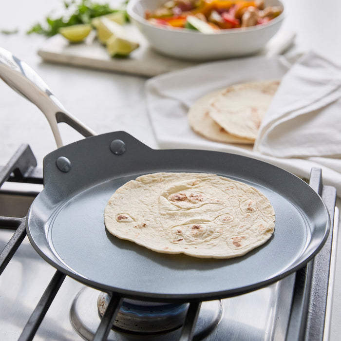 Tortilla on a grey ceramic 9.5 crepe pan on a gas stove, with toppings and tortillas beside the pan.