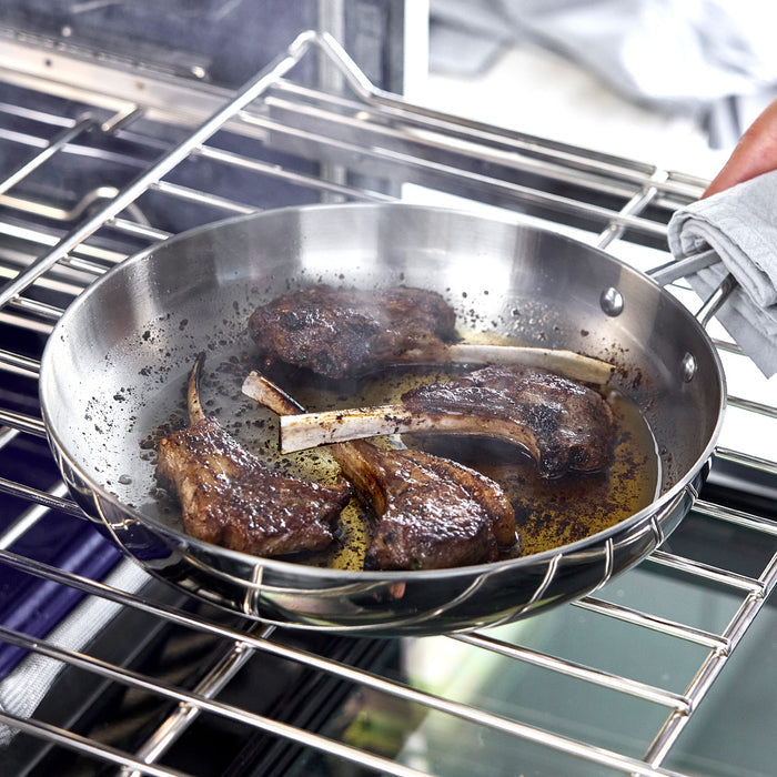Four lamb chops cooking in a stainless steel frypan, held by a person inside an oven.