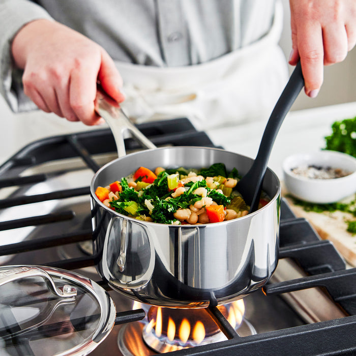 Person stirring soup in stainless steel saucepan with lid on a gas stove.