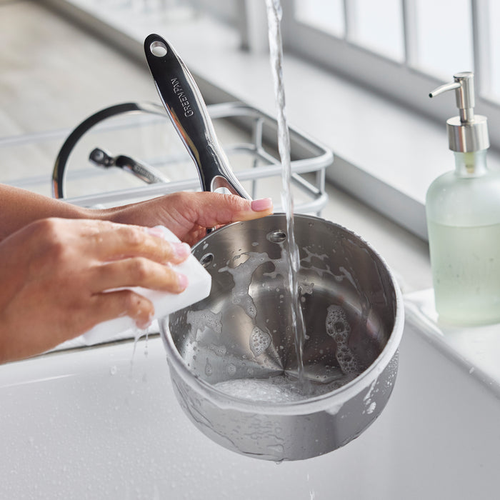 Washing a stainless steel saucepan with lid under running water in a kitchen sink.