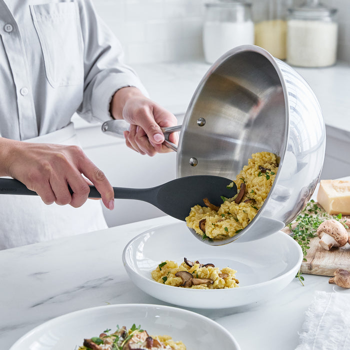 Person serving pasta with mushrooms from a chef's pan into a white bowl on a marble countertop.