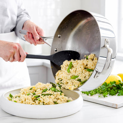 Rice and green vegetables being served from a stainless steel sauté pan into a white bowl.