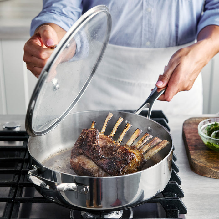 Person in light blue shirt and white apron lifts lid from 5-quart Valencia Pro sauté pan with seared lamb inside.
