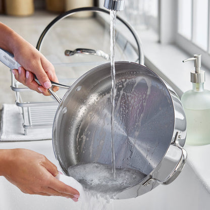A soapy stainless steel sauté pan with lid is rinsed under running water in a kitchen sink.